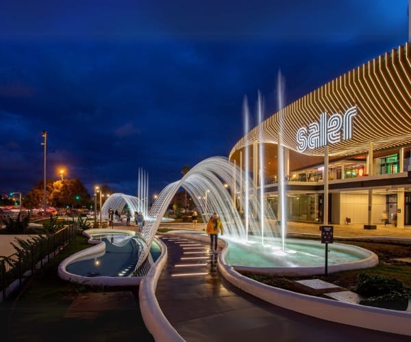 Illuminated arcing jets over a plaza pool outside a retail center at night