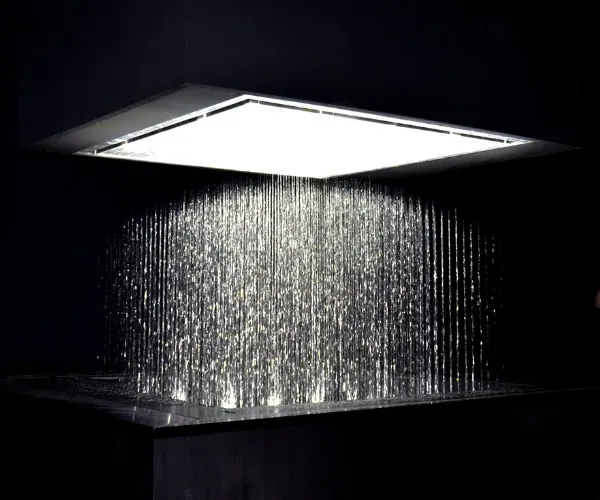 Rain curtain of lit streams falling from a square stainless manifold into a dark basin