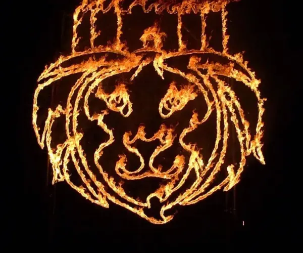 Steel lion-mask sculpture outlined in flame against a night sky