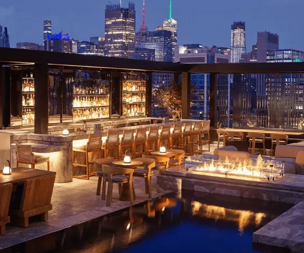 Linear fire trough on a rooftop bar terrace beside a lit plunge pool, city skyline at dusk