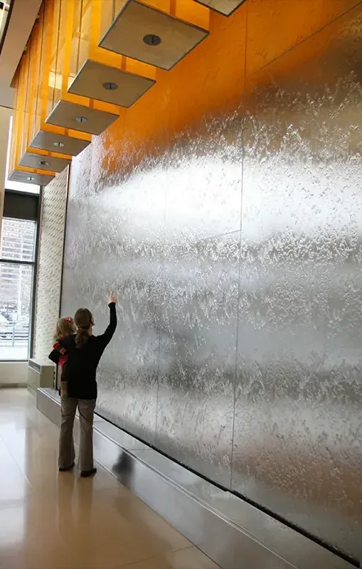 A parent and child look up at a full-height stainless waterwall running a continuous sheet of water in a warm-lit lobby